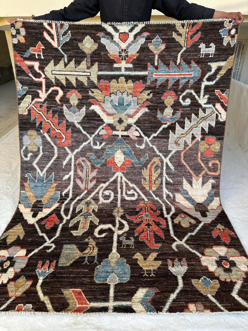 K&ouml;nnte beinhalten: Ein rechteckiger Teppich mit dunkelbraunem Hintergrund und einem symmetrischen Muster aus stilisierten Blumenmotiven, Bl&auml;ttern und kleinen Tierfiguren in Rot-, Blau-, Beige- und Wei&szlig;t&ouml;nen, abgeschlossen mit einer kurzen Franse am oberen Rand.