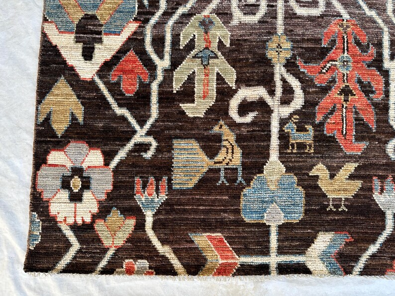 K&ouml;nnte beinhalten: Ein dunkelbrauner Teppich mit einem gewebten Muster aus stilisierten Blumen, V&ouml;geln und geometrischen Formen in Rot-, Blau-, Beige- und Wei&szlig;t&ouml;nen. Das Design enth&auml;lt komplexe, verzweigte Linien, die die verschiedenen Blumen- und Tiermotive verbinden.