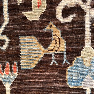 K&ouml;nnte beinhalten: Ein dunkelbrauner Webteppich mit einem wiederkehrenden Muster aus stilisierten V&ouml;geln, Blumenmotiven und geometrischen Formen in Hellblau, Beige und Rot. Die Textur ist dick und faserig, mit durchgehend sichtbaren horizontalen Linien.