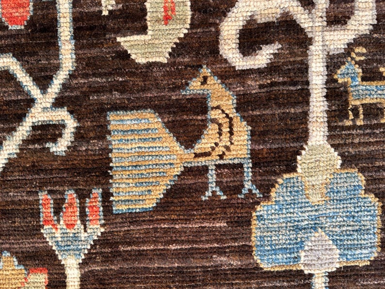 K&ouml;nnte beinhalten: Ein dunkelbrauner Webteppich mit einem wiederkehrenden Muster aus stilisierten V&ouml;geln, Blumenmotiven und geometrischen Formen in Hellblau, Beige und Rot. Die Textur ist dick und faserig, mit durchgehend sichtbaren horizontalen Linien.