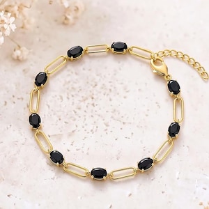 Könnte beinhalten: Ein goldfarbenes Kettenarmband mit einer Reihe ovaler schwarzer Steine, die in längliche Metallglieder eingefasst sind. Das Armband verfügt über eine zarte Verlängerungskette mit einem Karabinerverschluss und wirkt als strukturiertes Schmuckstück.