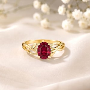 Puede incluir: Un anillo de oro con una piedra preciosa de color rojo intenso de corte ovalado sujeta por cuatro garras. La banda presenta un diseño delicado de enredadera entrelazada que se ramifica desde el engaste central, creando una estructura fluida.