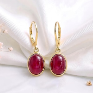 Puede incluir: Un par de pendientes de oro con cierre de palanca que presentan piedras cabujón ovaladas de color rojo intenso. Los pendientes tienen un engaste de metal dorado pulido que asegura las gemas rojas lisas y redondeadas.