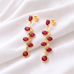 May include: A pair of gold-tone drop earrings featuring a zigzag metal bar design. Each earring is adorned with six oval-shaped, deep red faceted stones arranged along the length of the bar, ending with a single round red stone at the base.