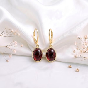 Peut inclure: Une paire de boucles d'oreilles en or avec fermoir levier, ornées de pierres précieuses grenat rouge foncé de forme ovale et lisse. Les boucles sont suspendues à une monture en métal doré poli qui maintient les pierres dans un design pendant.