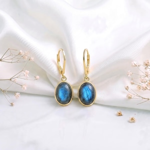 May include: A pair of gold leverback earrings featuring oval-shaped labradorite gemstones. Each earring displays a smooth, polished cabochon stone with a distinct blue iridescent flash set within a simple gold-toned metal bezel frame.