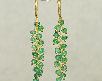 May include: A pair of gold-colored earrings with dangling strands of small, green gemstones. The earrings are suspended from a thin, light-colored branch. The gemstones are clustered together, creating a cascading effect.