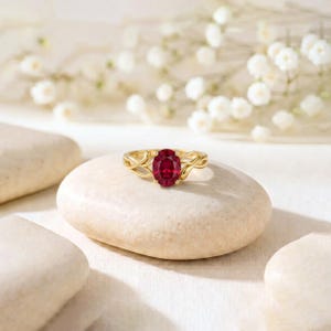 K&ouml;nnte beinhalten: Ein Goldring mit einem oval geschliffenen, tiefroten Edelstein in einer zarten, verschlungenen Ringschiene. Der Ring liegt auf einem glatten, hellen Stein und pr&auml;sentiert die leuchtende Farbe des Edelsteins sowie die filigrane Metallarbeit.