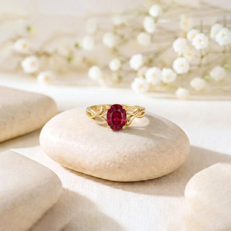 K&ouml;nnte beinhalten: Ein Goldring mit einem oval geschliffenen, tiefroten Edelstein in einer zarten, verschlungenen Ringschiene. Der Ring liegt auf einem glatten, hellen Stein und pr&auml;sentiert die leuchtende Farbe des Edelsteins sowie die filigrane Metallarbeit.