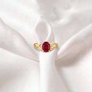 K&ouml;nnte beinhalten: Ein Goldring mit einem zentralen, oval geschliffenen, tiefroten Rubin in einer Vier-Krappen-Fassung. Die Ringschiene ist mit einem filigranen, ineinander verschlungenen, rankenartigen Design gestaltet, das vom Mittelstein ausgeht und eine organische Struktur bildet.
