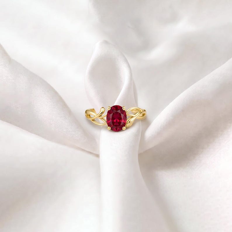 K&ouml;nnte beinhalten: Ein Goldring mit einem zentralen, oval geschliffenen, tiefroten Rubin in einer Vier-Krappen-Fassung. Die Ringschiene ist mit einem filigranen, ineinander verschlungenen, rankenartigen Design gestaltet, das vom Mittelstein ausgeht und eine organische Struktur bildet.