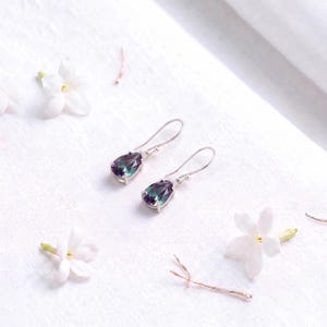 May include: A pair of silver drop earrings featuring faceted, pear-shaped stones that display a color-shifting gradient of deep purple, blue, and green hues. The earrings are suspended from simple silver hooks and are set in a polished metal frame.