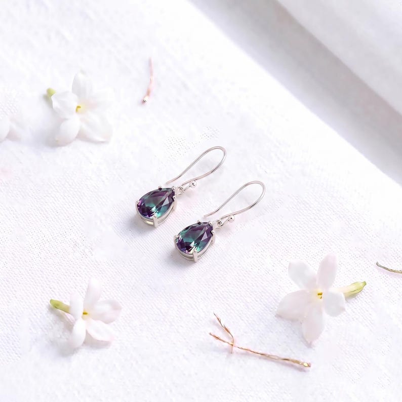 May include: A pair of silver drop earrings featuring faceted, pear-shaped stones that display a color-shifting gradient of deep purple, blue, and green hues. The earrings are suspended from simple silver hooks and are set in a polished metal frame.