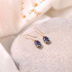 May include: A pair of gold drop earrings featuring pear-shaped, faceted stones that display a color-shifting gradient of deep purple, blue, and teal. The stones are secured in gold-toned metal settings with simple, curved gold ear wires.