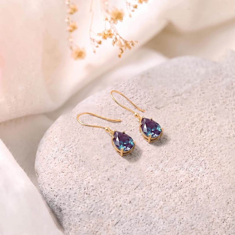 May include: A pair of gold drop earrings featuring pear-shaped, faceted stones that display a color-shifting gradient of deep purple, blue, and teal. The stones are secured in gold-toned metal settings with simple, curved gold ear wires.