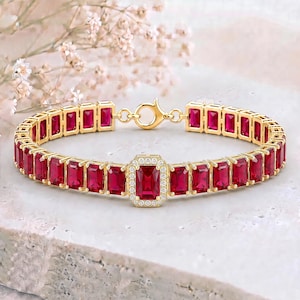 May include: A gold bracelet featuring a series of rectangular, emerald-cut red stones set in metal frames. The centre stone is surrounded by a halo of small white stones. The bracelet includes a circular clasp mechanism for secure fastening.
