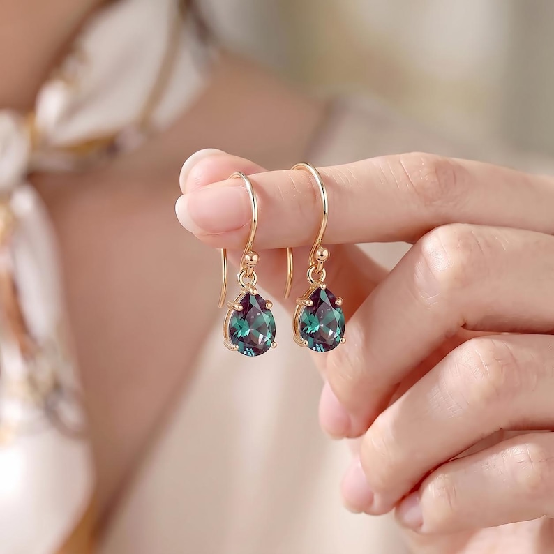 May include: A pair of gold-toned drop earrings featuring pear-shaped, faceted stones that transition between deep teal and purple hues. The earrings are suspended from simple curved gold-toned hooks, showcasing a polished metal finish.