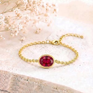 May include: A gold chain bracelet featuring a single oval-cut deep red gemstone set in a smooth gold bezel. The bracelet includes a delicate link chain and a small lobster claw clasp, showcasing a vibrant crimson stone as the central focal point.