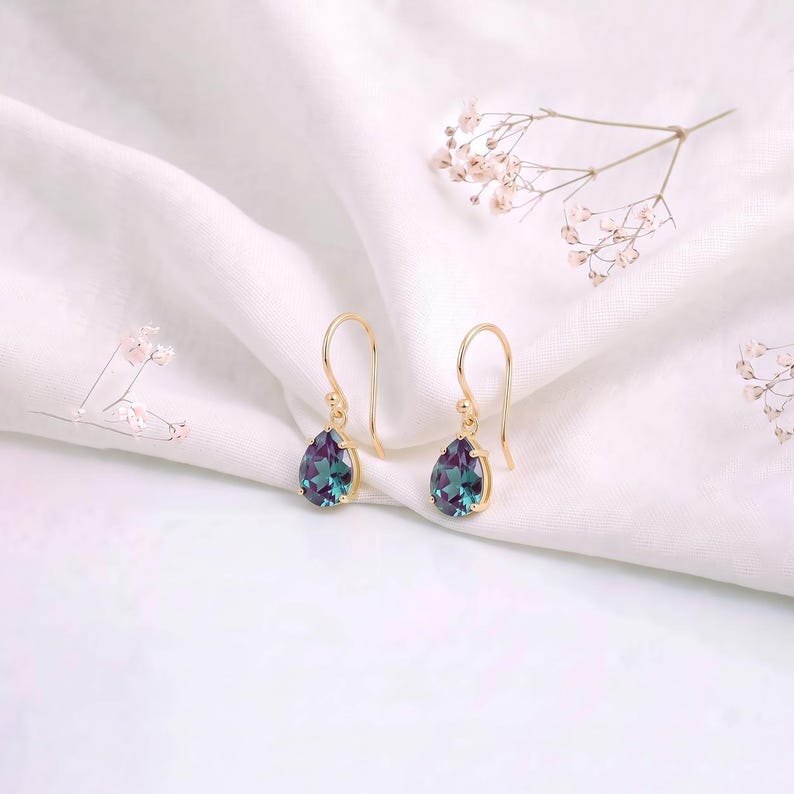May include: A pair of gold drop earrings featuring pear-shaped, color-changing stones that shift between deep purple and teal hues. The earrings are suspended from simple gold hooks, showcasing a faceted cut that reflects light from multiple angles.