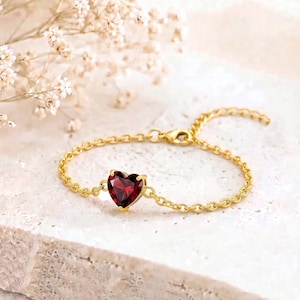 Peut inclure: Un bracelet à chaîne fine en or orné d'une pierre précieuse rouge unique en forme de cœur à facettes. Le bracelet est composé de maillons circulaires délicats et comprend un petit fermoir mousqueton pour une fixation sécurisée au poignet.