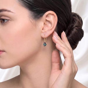 May include: A gold drop earring featuring a small, pear-shaped gemstone with iridescent hues of blue, green, and purple. The earring hangs from a simple gold hook wire, showcasing a delicate design that catches the light with its multifaceted surface.