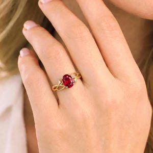 K&ouml;nnte beinhalten: Ein goldener Ring mit einem zentralen, oval geschliffenen, tiefroten Edelstein, der von vier Krappen gehalten wird. Die Ringschiene ist mit einem filigranen, rankenartigen Design gestaltet, das in die Fassung &uuml;bergeht und gl&auml;nzt.