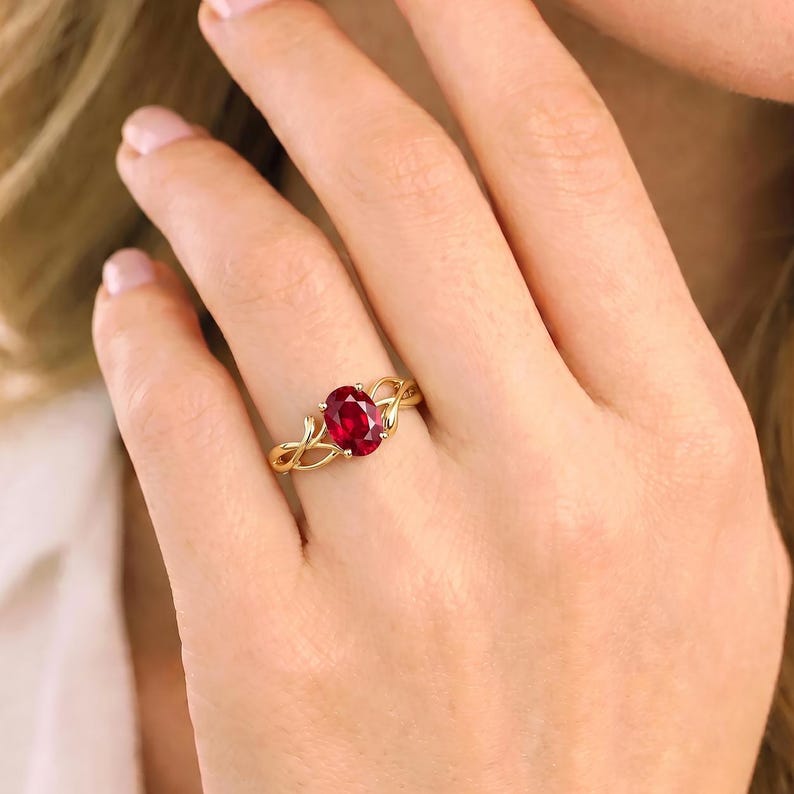 K&ouml;nnte beinhalten: Ein goldener Ring mit einem zentralen, oval geschliffenen, tiefroten Edelstein, der von vier Krappen gehalten wird. Die Ringschiene ist mit einem filigranen, rankenartigen Design gestaltet, das in die Fassung &uuml;bergeht und gl&auml;nzt.