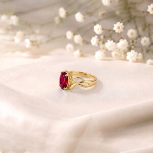 K&ouml;nnte beinhalten: Ein goldener Ring mit einem leuchtend roten Edelstein im Ovalschliff, der von vier Krappen gehalten wird. Die Ringschiene ist poliert und zeigt ein filigranes, durchbrochenes &Uuml;berkreuz-Design, das sich nach hinten verj&uuml;ngt.
