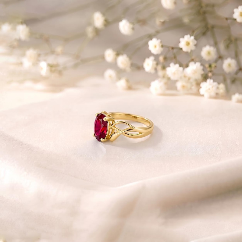 K&ouml;nnte beinhalten: Ein goldener Ring mit einem leuchtend roten Edelstein im Ovalschliff, der von vier Krappen gehalten wird. Die Ringschiene ist poliert und zeigt ein filigranes, durchbrochenes &Uuml;berkreuz-Design, das sich nach hinten verj&uuml;ngt.
