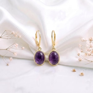 May include: A pair of gold-toned leverback earrings featuring oval-shaped, deep purple amethyst cabochon stones. The earrings have a polished metal setting that secures the smooth, rounded purple gemstones in a simple, classic drop design.