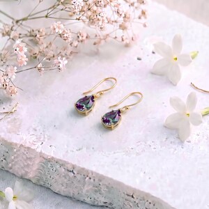 May include: A pair of gold drop earrings featuring pear-shaped, iridescent, multi-colored gemstones. The stones display shifting hues of purple, green, and blue, suspended from simple gold hooks. The earrings are displayed on a textured white surface.
