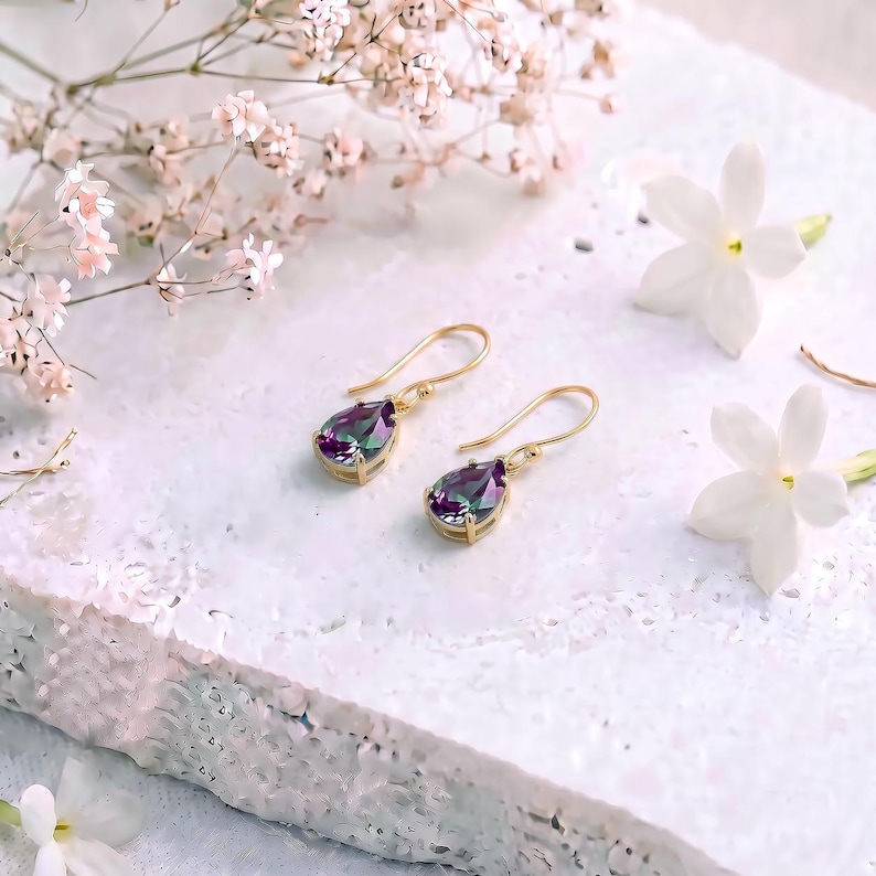 May include: A pair of gold drop earrings featuring pear-shaped, iridescent, multi-colored gemstones. The stones display shifting hues of purple, green, and blue, suspended from simple gold hooks. The earrings are displayed on a textured white surface.
