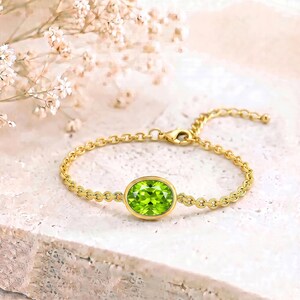 May include: A gold-toned chain bracelet featuring a single oval-cut, vibrant lime-green gemstone set in a smooth bezel. The bracelet includes a delicate link chain and a small lobster claw clasp, showcasing a bright and polished metallic finish.
