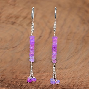 May include: A pair of long drop earrings featuring silver hooks and chains. Each earring displays a vertical row of small, round, iridescent purple beads, with two additional purple beads dangling from the bottom of each silver chain segment.