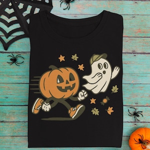 Puede incluir: Camiseta negra con un diseño de calabaza y fantasma de dibujos animados, ambos corriendo. El diseño incluye estrellas, hojas y caramelos. Prenda con temática de Halloween.