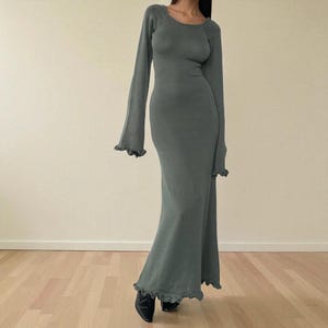Peut inclure: Une longue robe vert sauge avec des manches longues et une encolure arrondie. Les manches s'&eacute;vasent aux poignets, et l'ourlet de la robe a &eacute;galement un bord &agrave; volants. La robe est faite d'une mati&egrave;re douce et moulante.