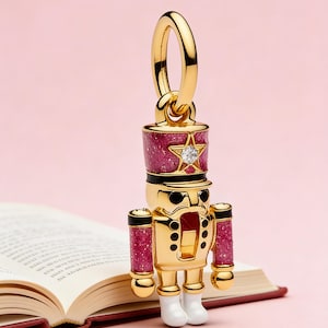 May include: A gold-colored nutcracker charm with a red and glittery hat and arms, a star-shaped gem, and white boots. The charm is hanging from a gold ring. The background is a pink surface and an open book.