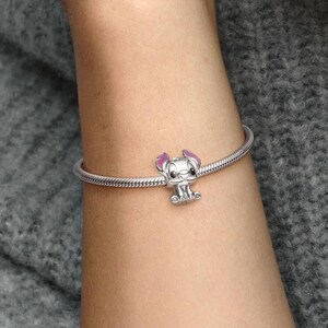 May include: A silver bracelet with a charm featuring a cartoon character with pink ears and a small, detailed face. The bracelet is worn on a person's arm, against a gray background.