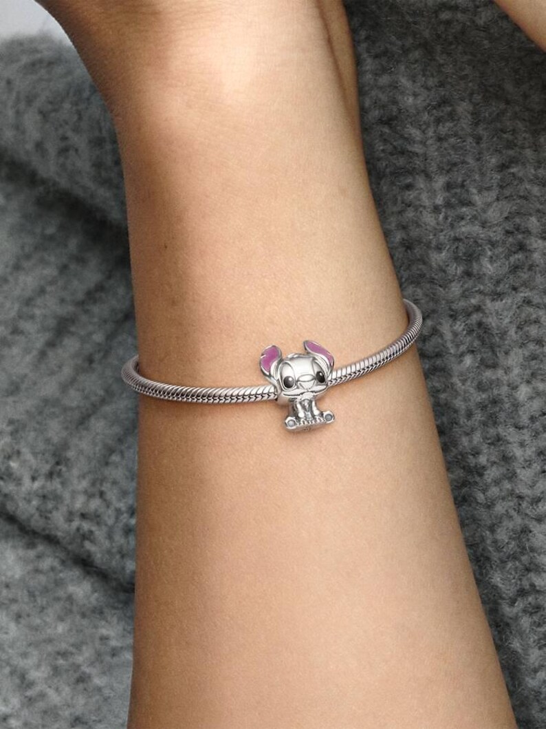 May include: A silver bracelet with a charm featuring a cartoon character with pink ears and a small, detailed face. The bracelet is worn on a person's arm, against a gray background.
