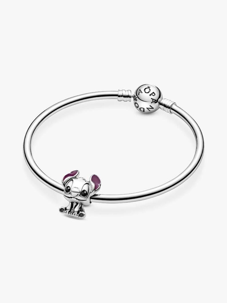 May include: A silver bracelet with a charm featuring a cartoon character with purple ears and accents. The charm is attached to a silver bangle-style bracelet. The bracelet has a round clasp with the brand name on it.