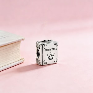 Puede incluir: Un charm de plata con forma de libro con las palabras "FAIRY TALE" y un diseño de corona. El charm tiene detalles intrincados y está sobre un fondo rosa suave.