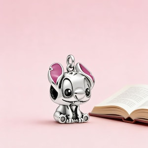 May include: A silver-colored charm in the shape of the Disney character Stitch, with pink accents on the ears. The charm is sitting on a pink background next to an open book.
