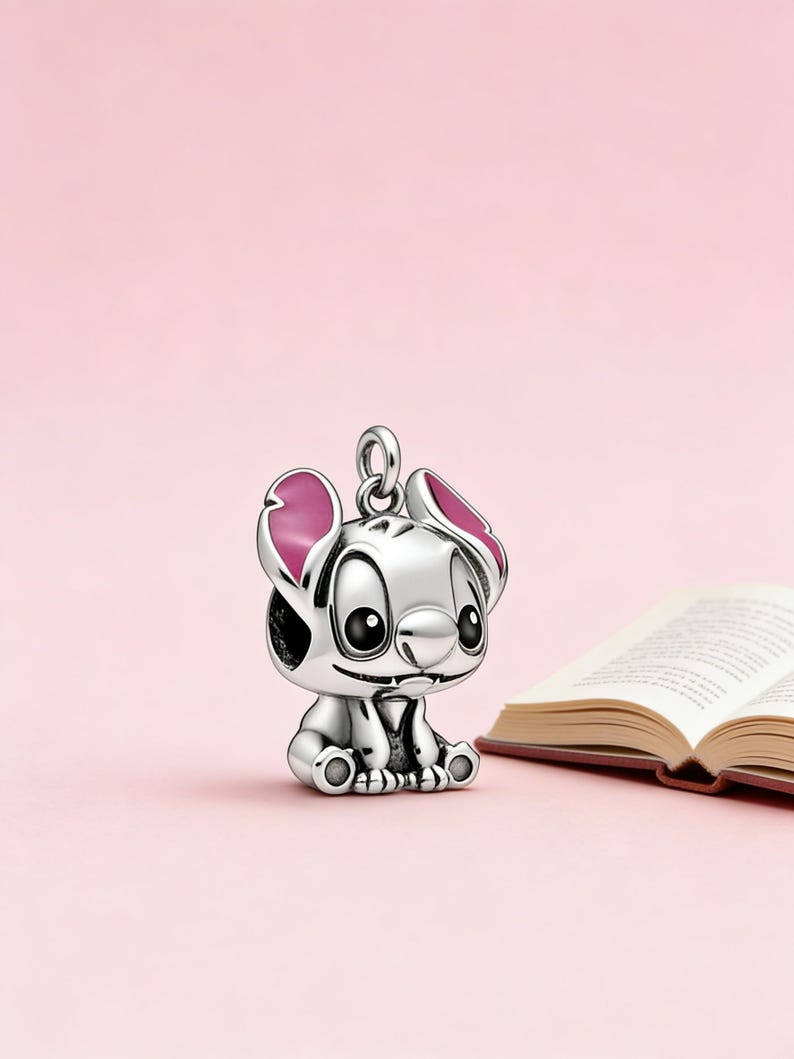 May include: A silver-colored charm in the shape of the Disney character Stitch, with pink accents on the ears. The charm is sitting on a pink background next to an open book.