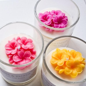 以下が含まれることがあります： 花柄デザインのガラス瓶入りキャンドル3つ。1つのキャンドルには明るいピンクの花、別のキャンドルには黄色の花、3つ目にはピンクの花があります。キャンドルには「ROSE & EMBER COZY AND WARM」と表示されています。