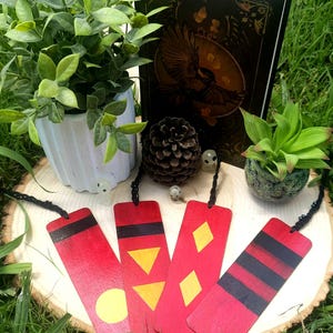 Puede incluir: Un conjunto de marcadores de libros de madera hechos a mano con diseños geométricos en amarillo y negro sobre un fondo rojo. Los marcadores se exhiben sobre una rodaja de madera con un libro, plantas y objetos decorativos.