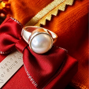 Puede incluir: Un anillo de plata con una perla blanca iridiscente, grande y redonda, engastada en un bisel de metal liso. El anillo está colocado sobre una caja de regalo roja con un lazo rojo y una etiqueta con el texto visible "Wishing you a very".