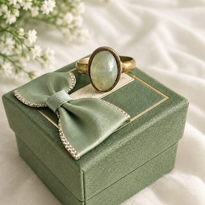 Puede incluir: Un anillo ovalado con una piedra de color verde pálido engastada en una banda de metal color latón. El anillo descansa sobre una caja de regalo cuadrada de color verde salvia con un lazo de satén a juego, borde brillante y etiqueta blanca.