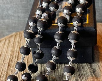 Puede incluir: Primer plano de un rosario con cuentas de madera oscura y detalles metálicos plateados. Las cuentas son esféricas y están conectadas por delicados eslabones metálicos. El rosario se muestra sobre una caja oscura y una superficie de madera.