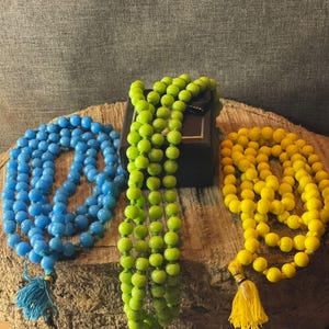 May include: Three beaded necklaces in blue, green, and yellow, each with a tassel. The necklaces are displayed on a wooden surface. The beads are round and evenly spaced, creating a simple, elegant design.