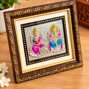 May include: A rectangular framed wall art piece featuring a silver-toned embossed metal plate with depictions of Lakshmi and Ganesha. The figures are accented with vibrant pink, blue, and gold colors. The frame is dark brown with gold floral detailing.